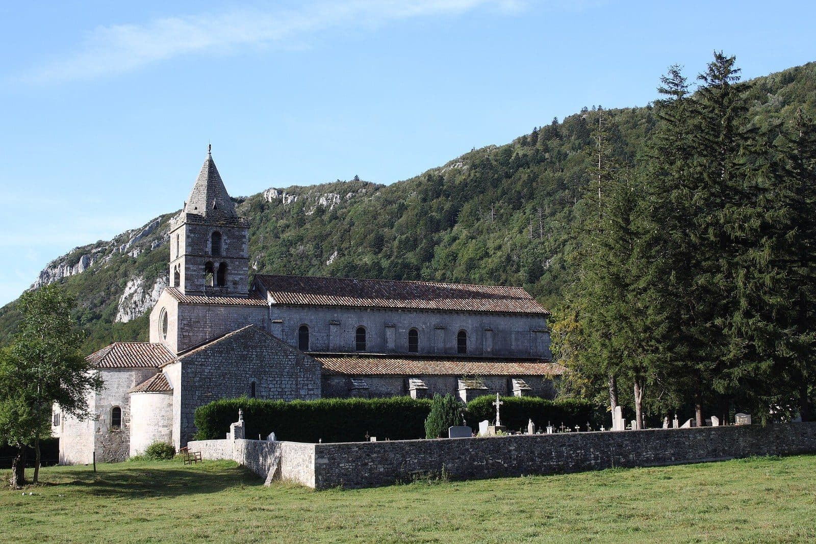 The Romanesque abbey church of Léoncel and its stone bell tower, surrounded by meadows at the foot of a wooded mountain.