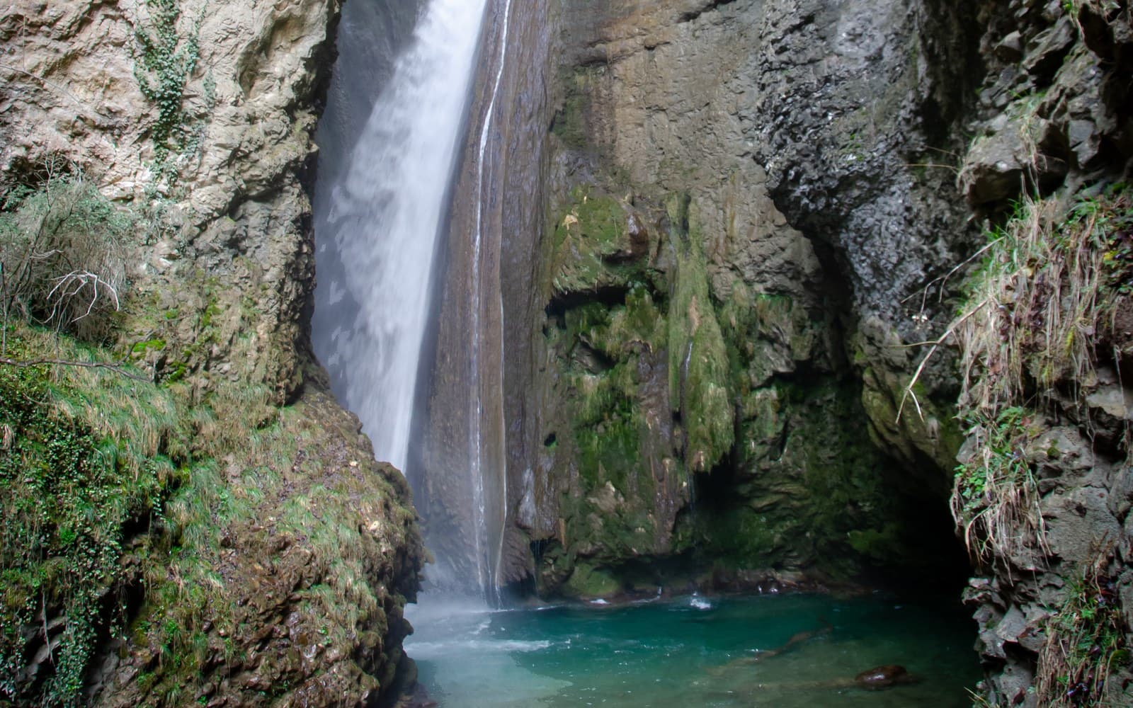The foot of the Druise waterfall: the white cascade plunges between mossy rock walls into a turquoise pool.