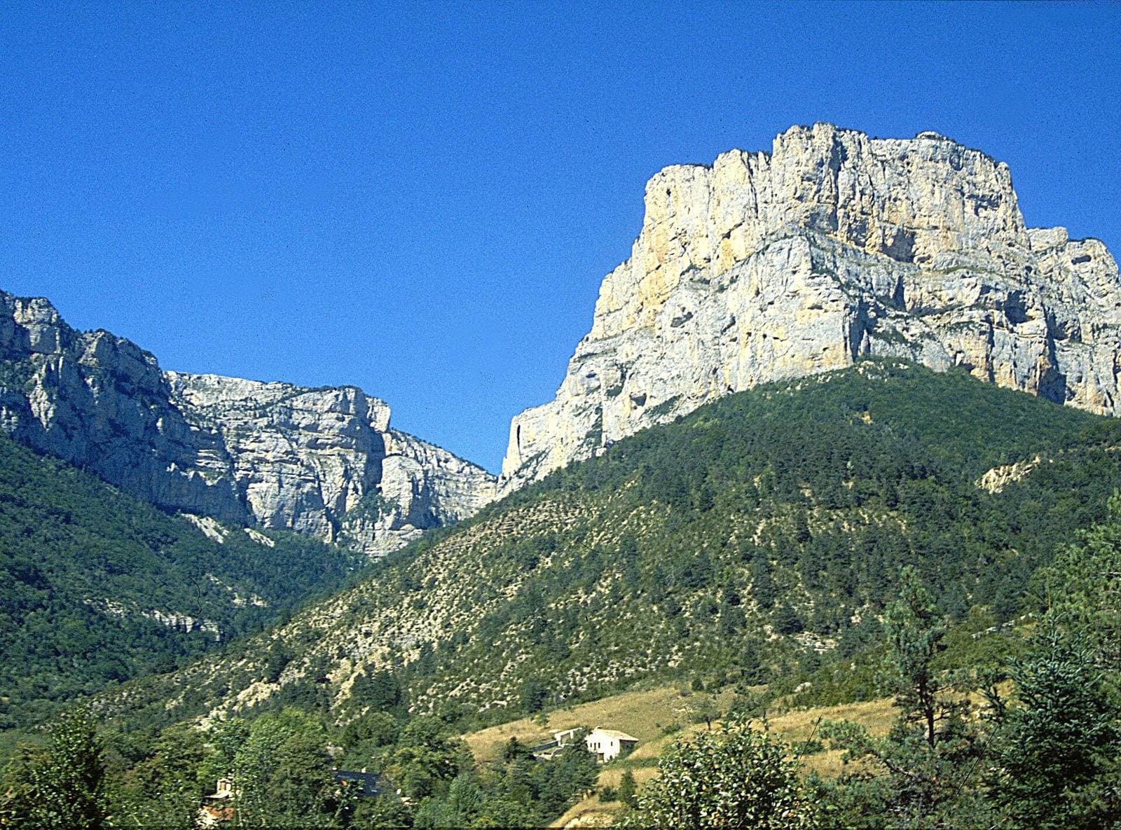 The limestone cliffs of the Cirque d’Archiane tower over wooded slopes and a few houses of the hamlet, beneath a deep blue sky.