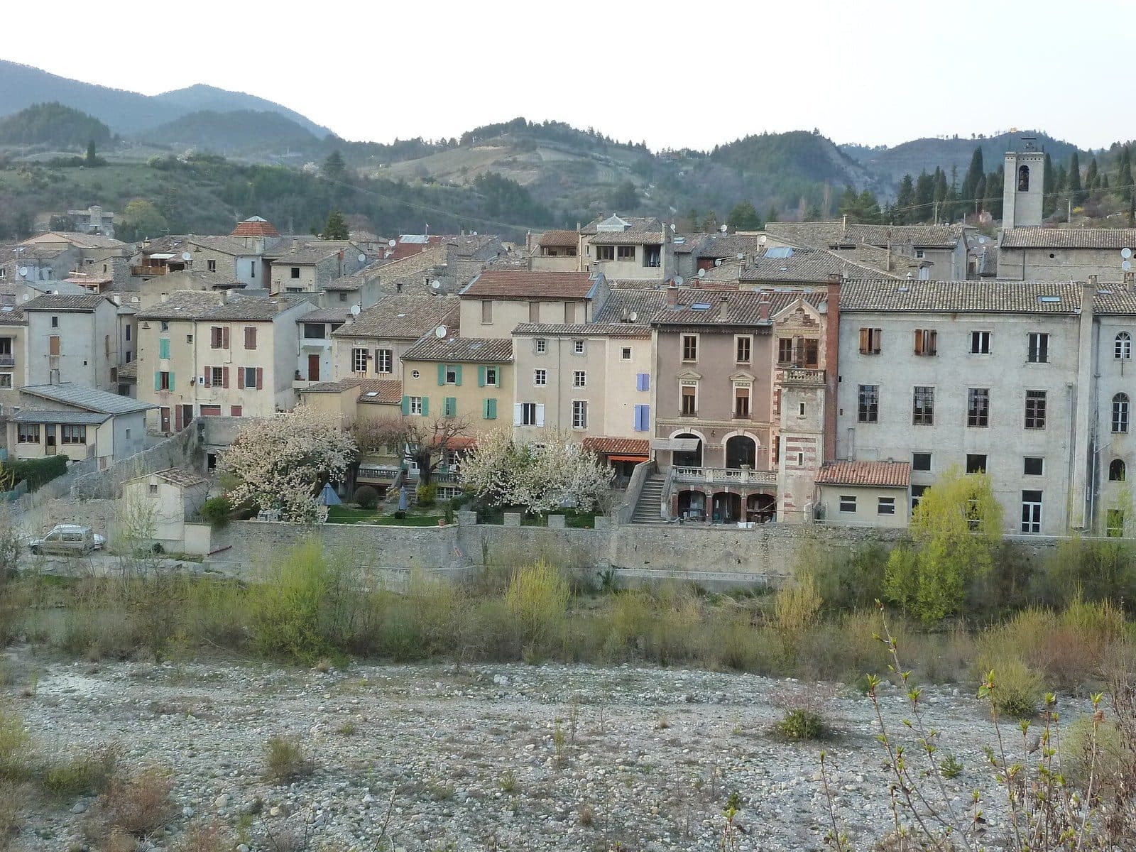 The old façades of Saillans beside the pebbly bed of the Drôme, with a bell tower and wooded hills in the distance.